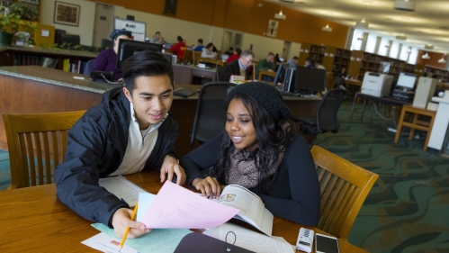 Two students look at papers and books in a campus library