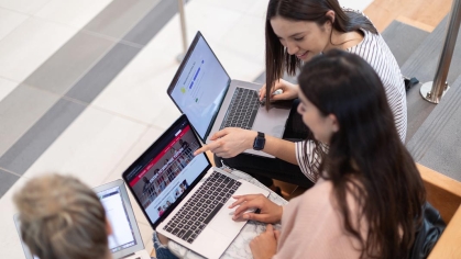 Three young female students, each with their own laptop, engaging together, pointing to screens