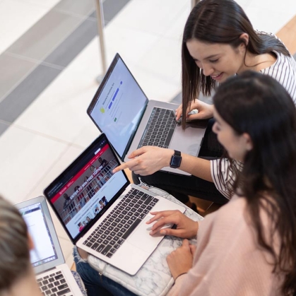 Three young female students, each with their own laptop, engaging together, pointing to screens