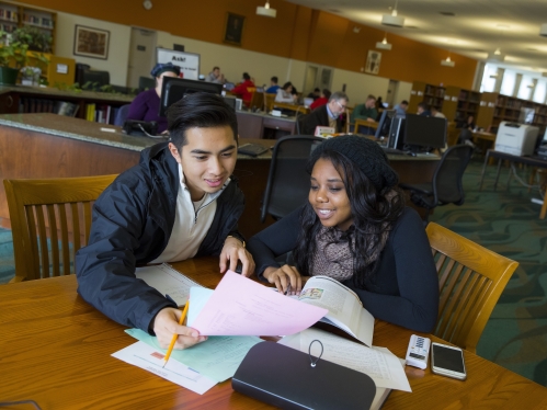 Two students look at papers and books in a campus library